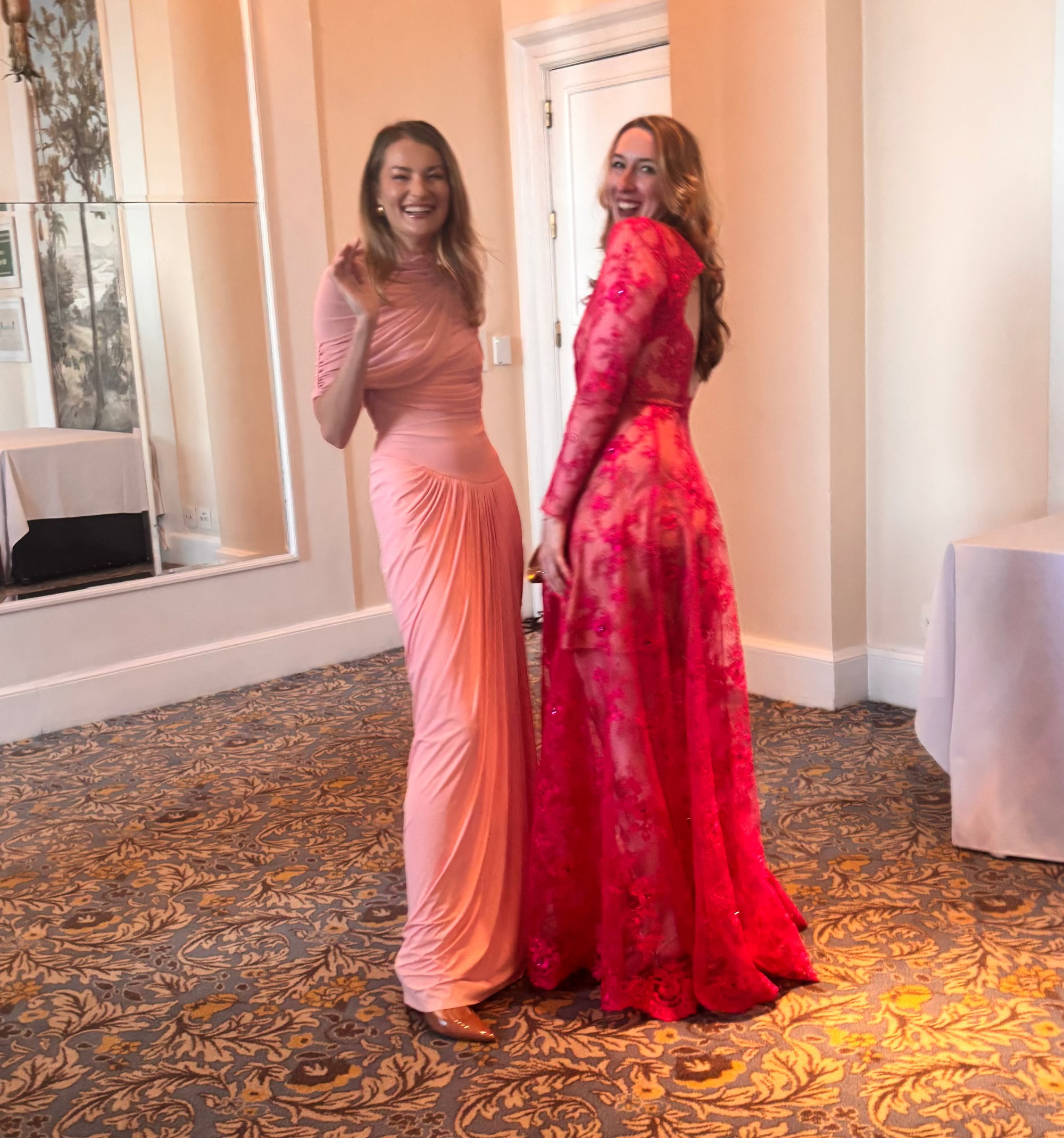 Two women in pink and red evening gowns standing in a room with patterned carpet.