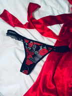 Black lace thong with red rose embroidery placed on a white bedspread alongside a flowing red satin ribbon.