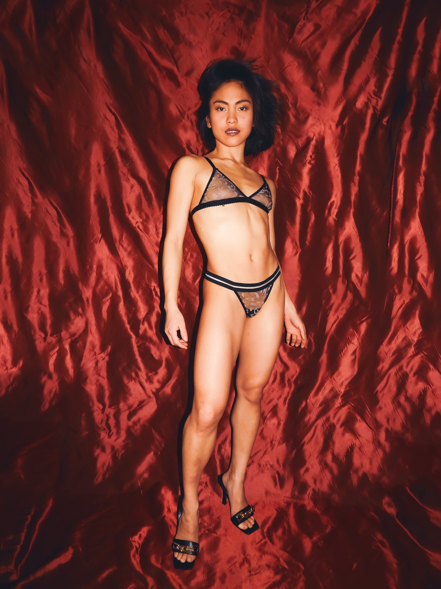 model in black mesh set and brown details standing with arms down against a red backdrop